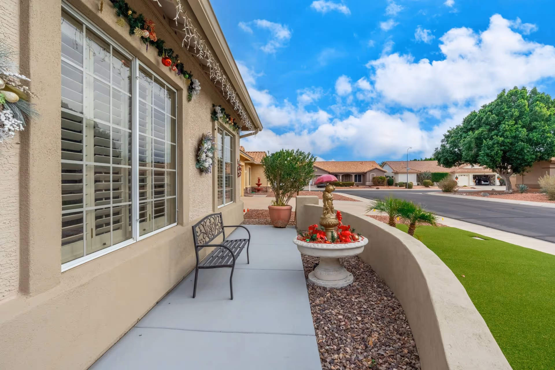 Outdoor walkway alongside a beige building with large windows decorated with holiday wreaths and garlands. A black metal bench is placed on the concrete path next to a decorative fountain with red flowers. The area is landscaped with rocks, small palm plants, and a green lawn. In the background, there are residential houses, a tree, and a partly cloudy blue sky.