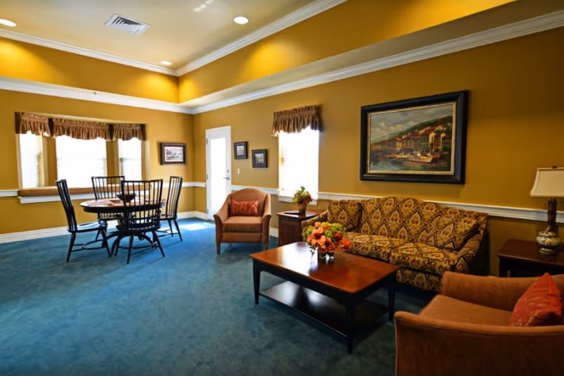 A cozy living room area with mustard yellow walls and blue carpet. The room features a patterned sofa, two armchairs with red cushions, a wooden coffee table with a flower arrangement, and a side table with a lamp. There is a round dining table with four chairs near the windows, which have brown valances. A framed painting hangs on the wall above the sofa.