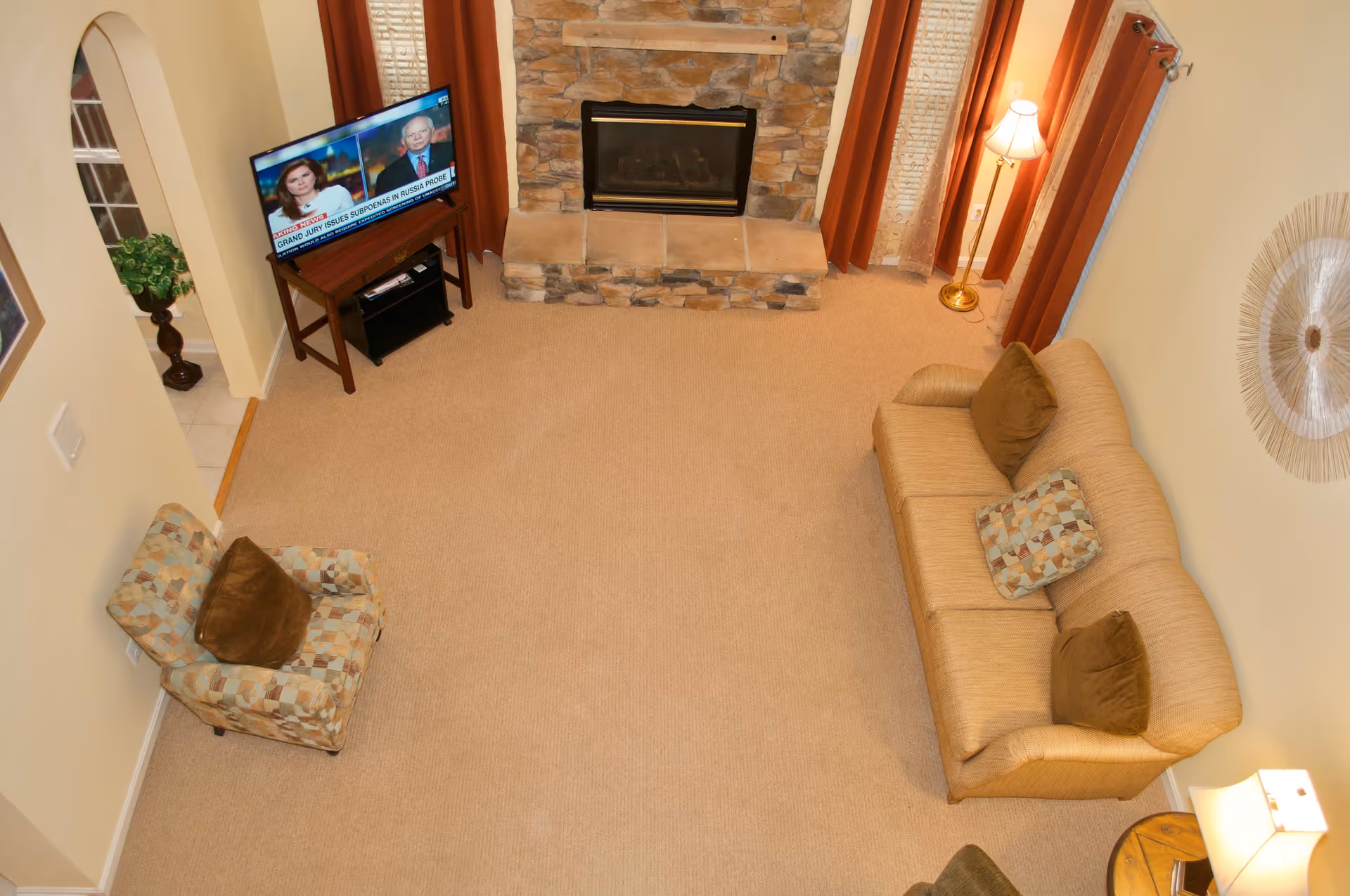 A cozy living room with beige carpet, a stone fireplace, a beige couch with brown and patterned pillows, a patterned armchair with a brown pillow, a floor lamp, a table lamp, and a TV on a stand showing a news broadcast.