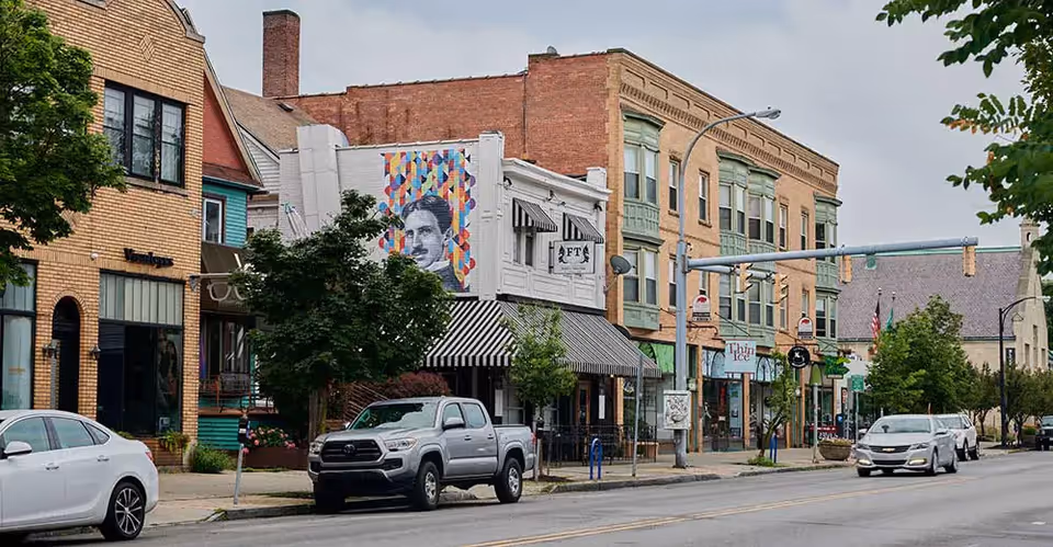 Street view of a small town commercial area with parked cars along the road. There are several buildings with storefronts, including one with a colorful mural of a man's face on the side. Trees line the sidewalk and traffic lights hang above the street.