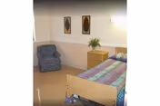 A patient bedroom with a bed, nightstand, plant, and an armchair.