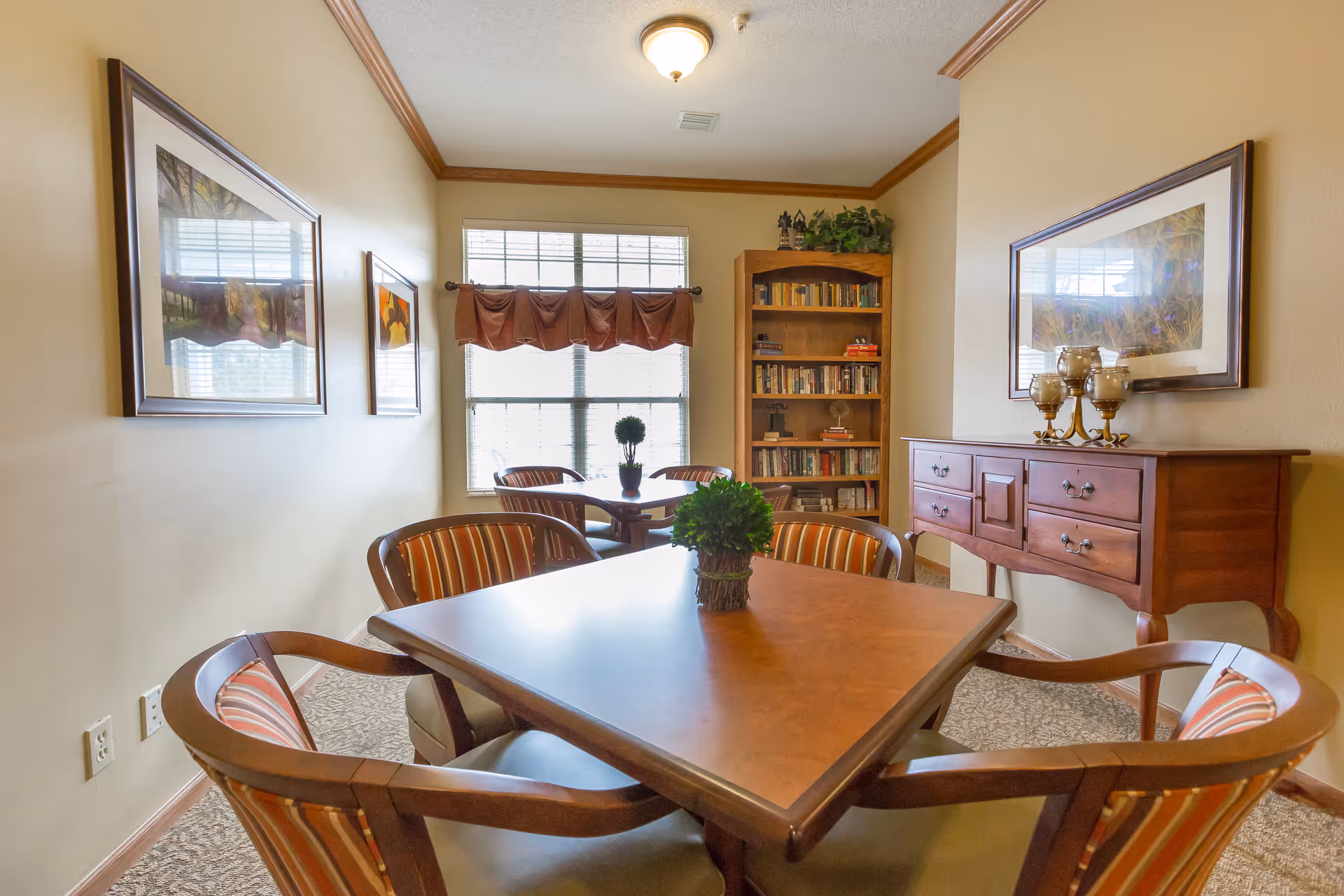 A cozy interior room with two wooden tables surrounded by striped cushioned chairs. Each table has a small green plant centerpiece. The room features beige walls with framed artwork, a wooden bookshelf filled with books, and a wooden sideboard with decorative candle holders. A window with blinds and a valance allows natural light to brighten the space.