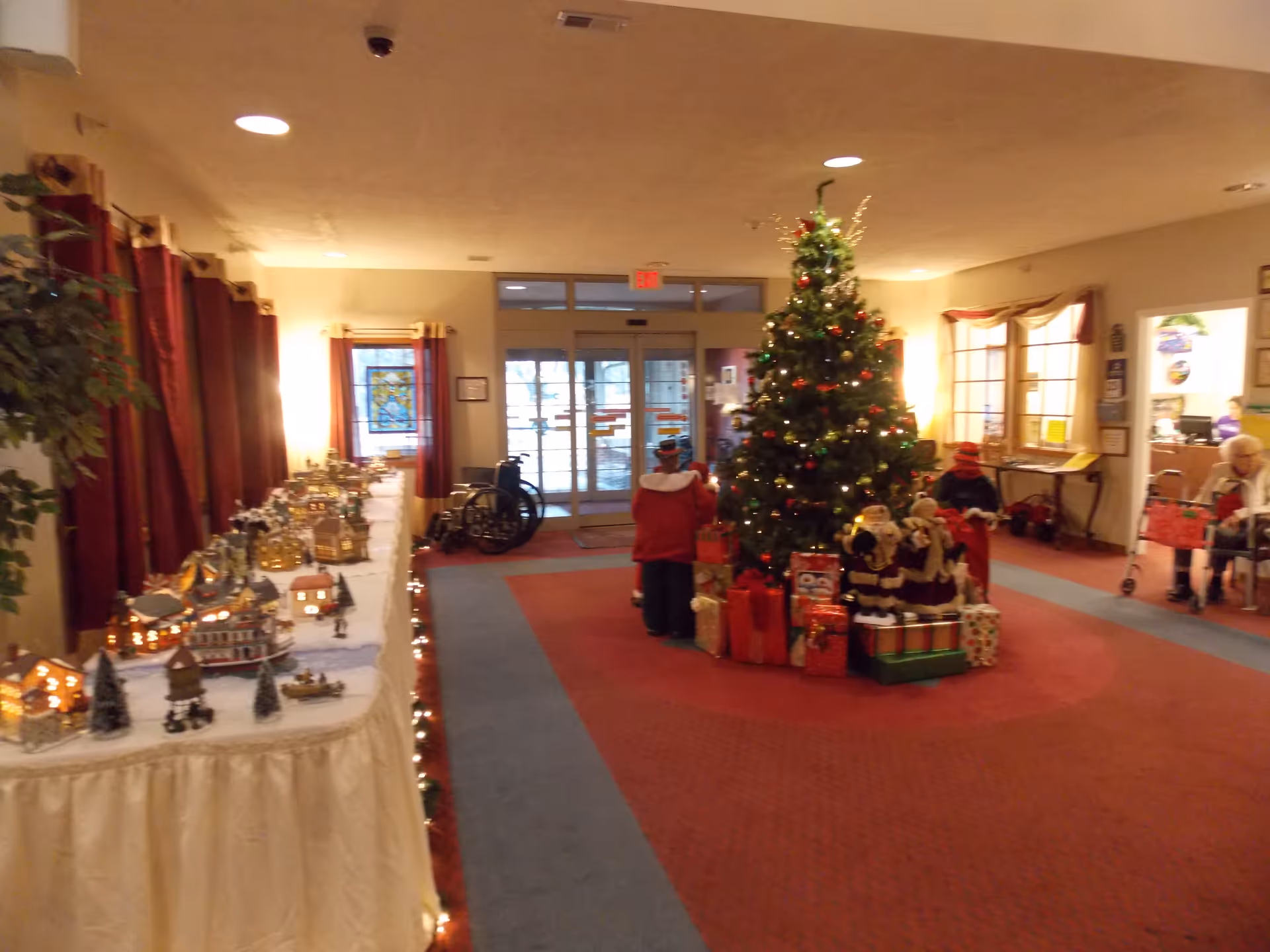 Interior of a senior living facility lobby decorated for Christmas with a large Christmas tree surrounded by wrapped presents and festive figurines. To the left, a long table displays a miniature holiday village with small houses and trees. There are wheelchairs and a couple of elderly residents visible near the entrance and seating area.