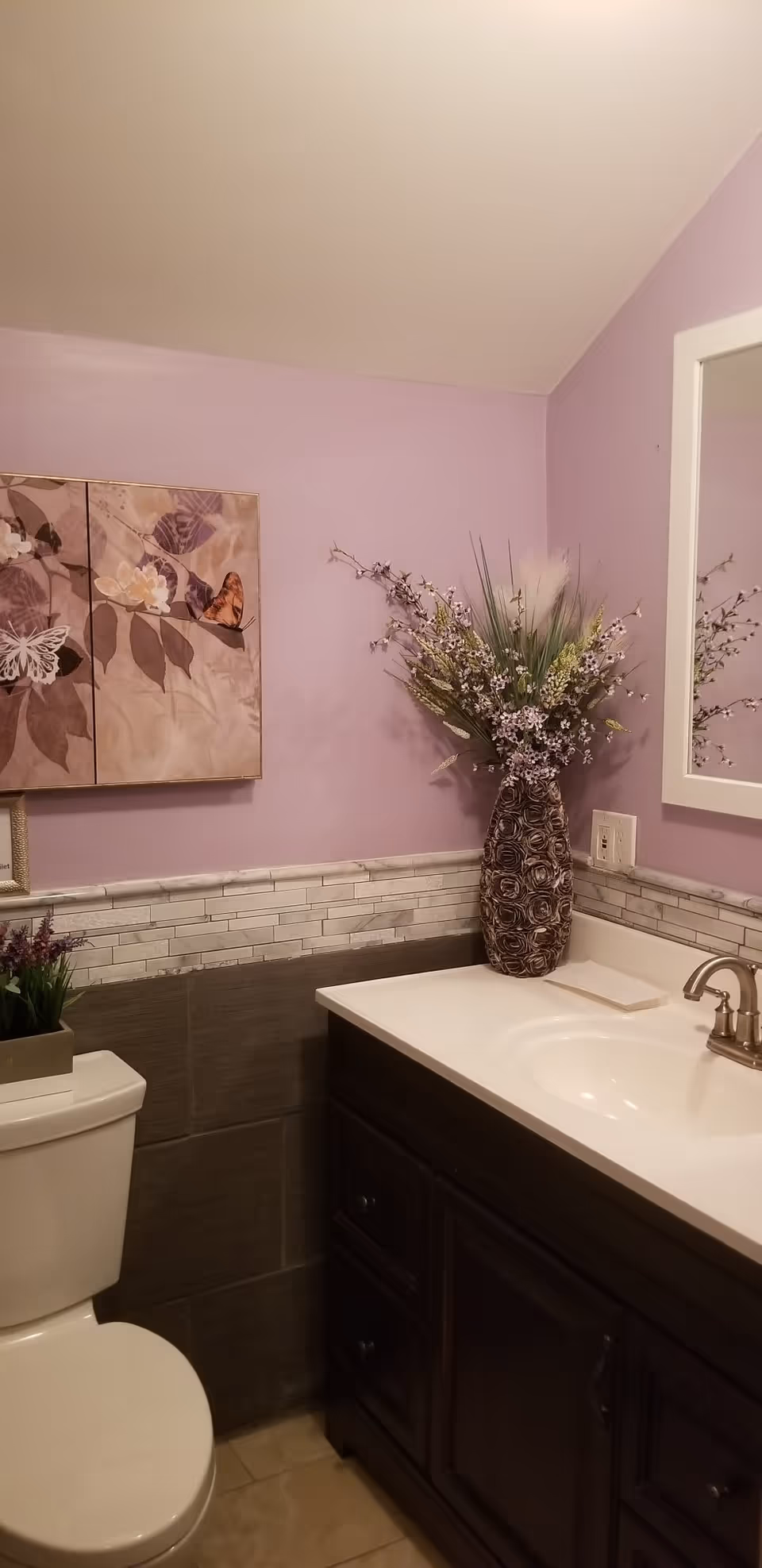 A bathroom with light purple walls and a white toilet. There is a dark wooden vanity with a white countertop and a silver faucet. On the countertop, there is a decorative vase with artificial flowers. Above the vanity is a mirror with a white frame. The wall has a decorative tile border halfway up, and there is a framed butterfly artwork on the wall above the toilet.
