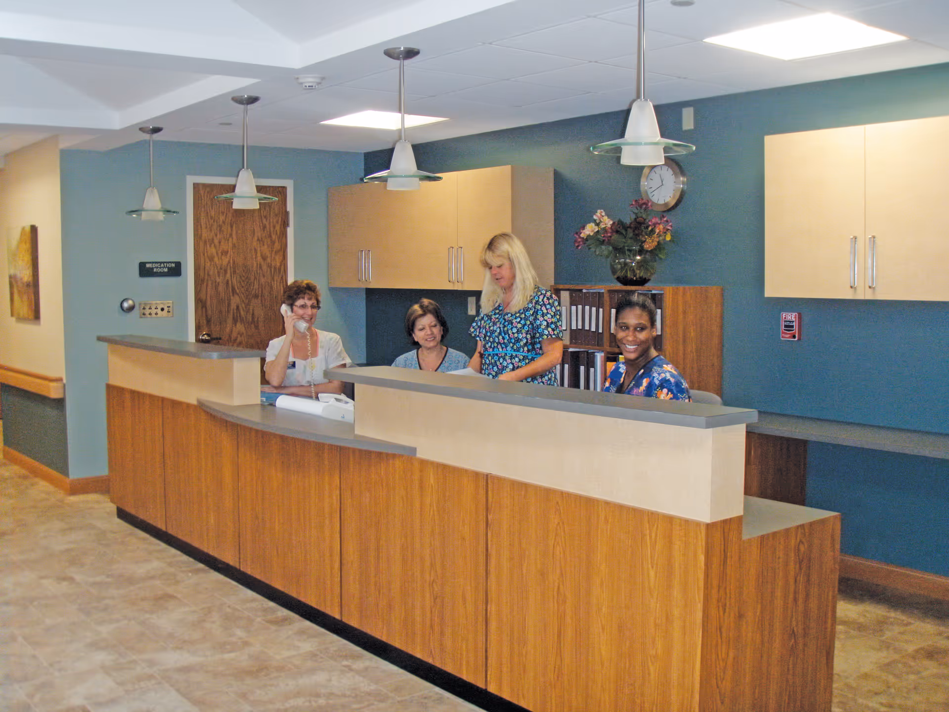 Reception area with four women behind a wooden front desk. One woman is on the phone, two are looking at documents, and one is smiling at the camera. The background includes blue walls, wooden cabinets, a clock, and a vase with flowers.