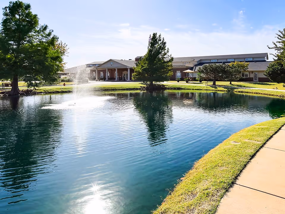 A landscaped pond with a central fountain in front of a low-rise senior living building and walkway.