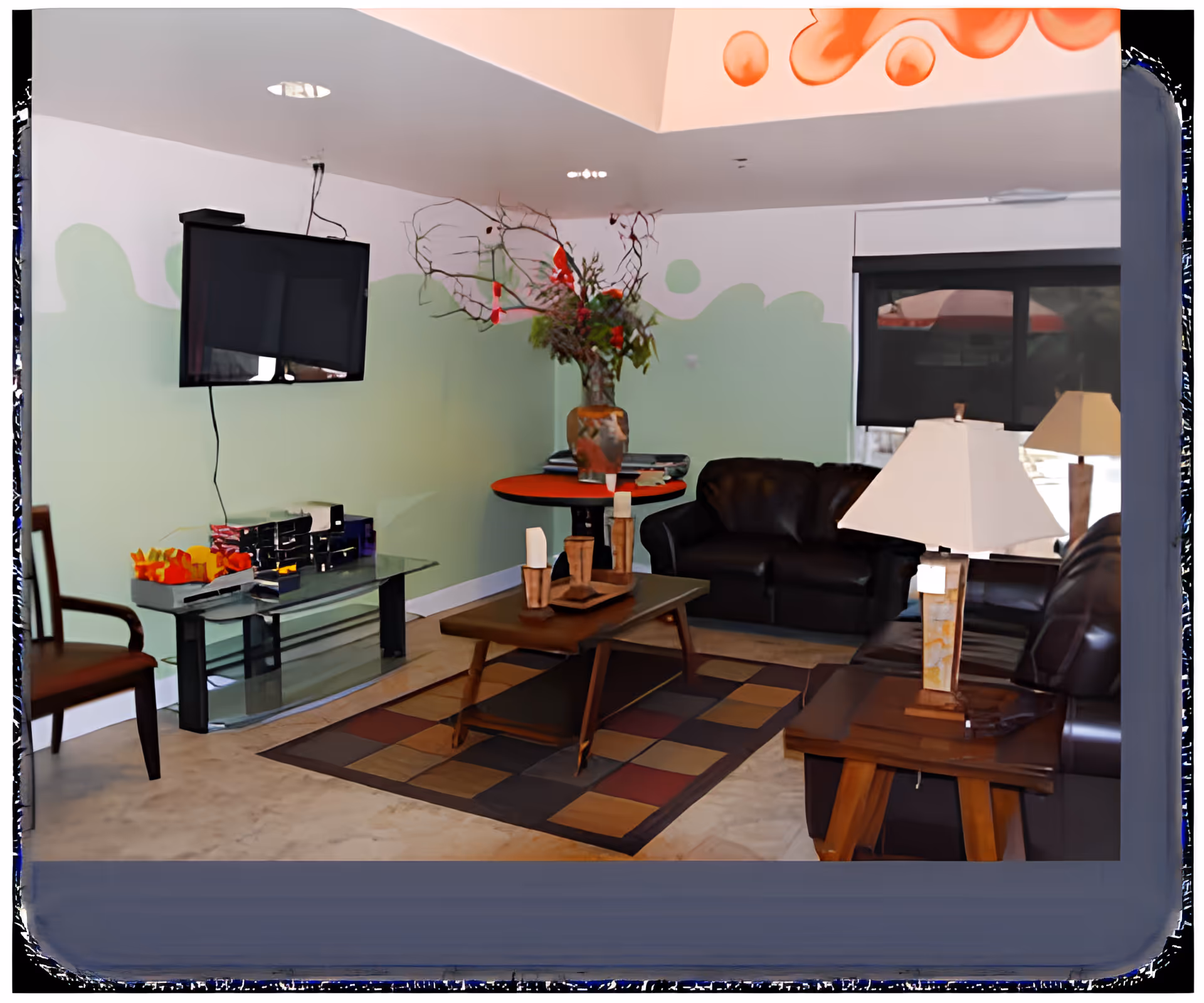 A cozy living room with two dark leather sofas, a wooden coffee table with decorative candles, a side table with a lamp, a wall-mounted flat-screen TV, and a small round table with a large vase of flowers. The walls are painted white with a green wavy pattern, and there is a window with a black roller shade partially drawn.