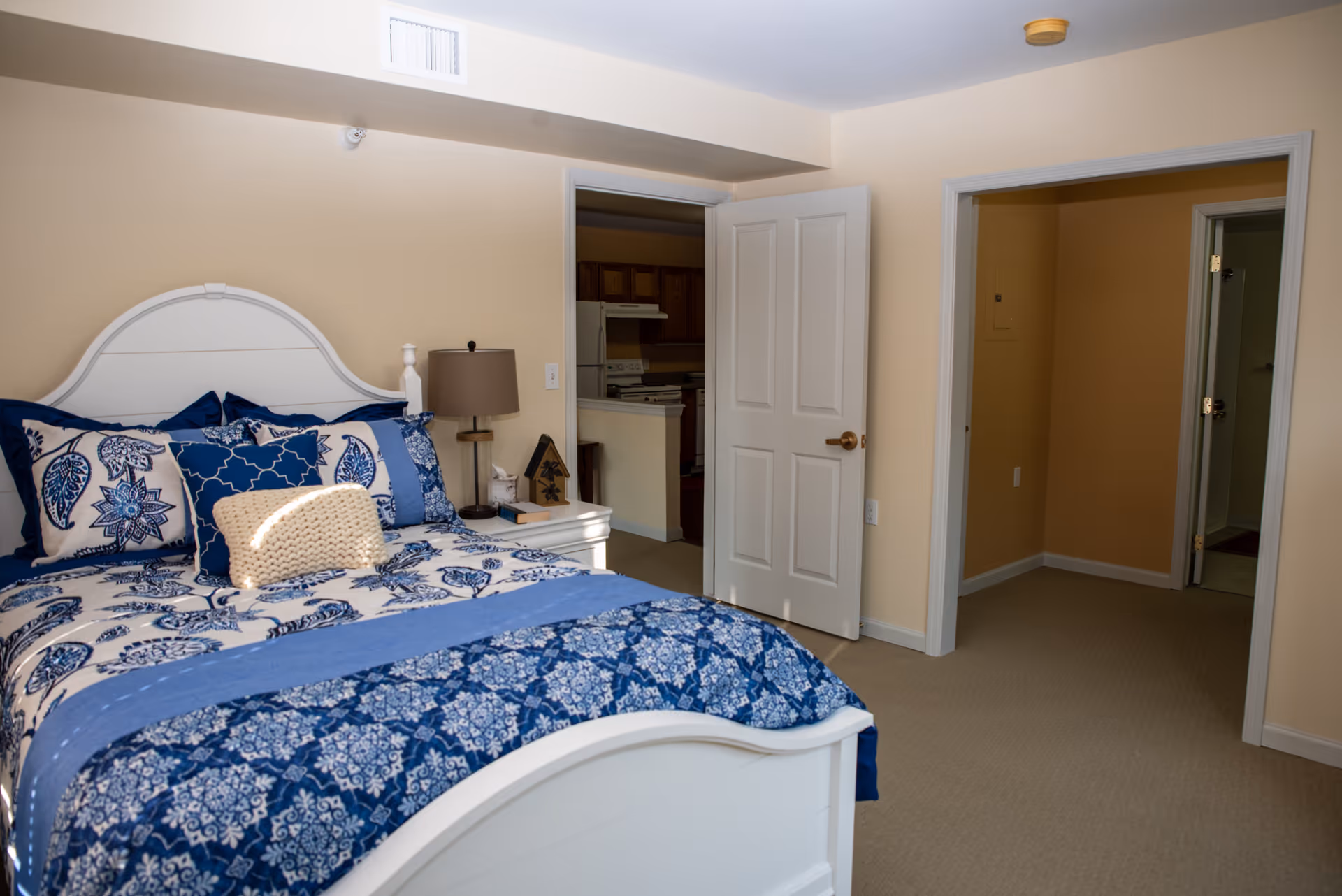 A bedroom with a white bed frame and blue patterned bedding, including multiple pillows. There is a nightstand with a lamp and decorative items next to the bed. The room has beige walls and carpeted floor. Two open doorways lead to other rooms, one showing a kitchen area with wooden cabinets and appliances.