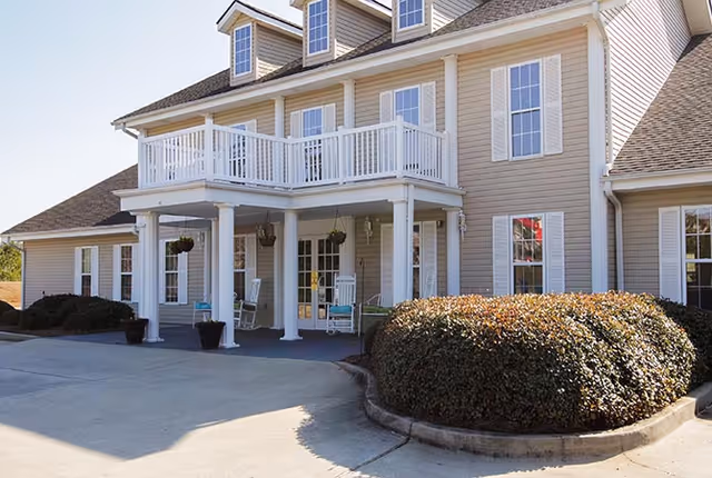 Front entrance of a two-story beige senior living building with white columns, a balcony, rocking chairs, and shrubbery.