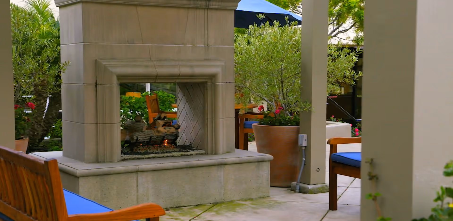 Outdoor patio seating area with a stone fireplace, wooden benches with blue cushions, and potted plants.
