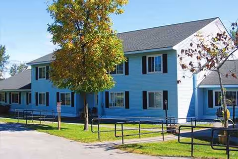 Blue two-story apartment building with black shutters, trees, and a ramped walkway.