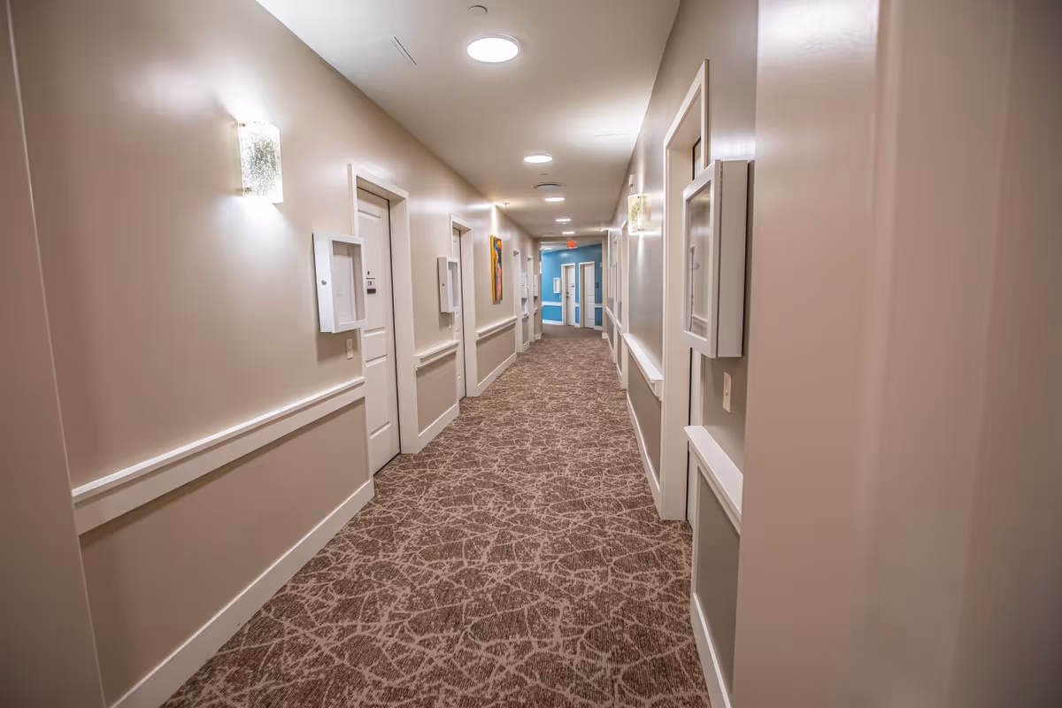 A well-lit hallway in a senior living facility with beige walls, patterned carpet, multiple closed white doors on both sides, wall-mounted light fixtures, and a blue accent wall at the far end.