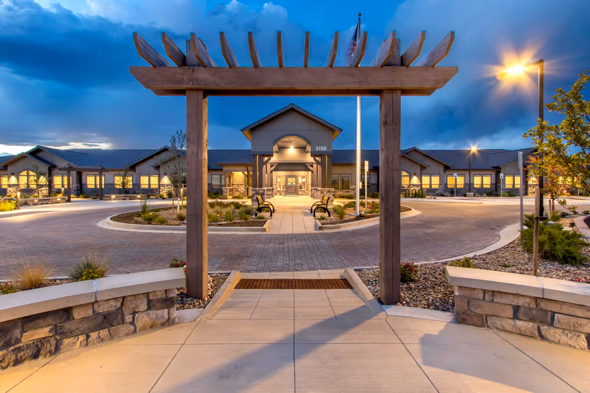 Evening view of the entrance to a single-story memory care facility with a wooden pergola in the foreground, a circular driveway, benches, landscaped plants, and the building illuminated with warm lights under a cloudy blue sky.