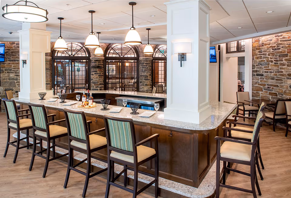 A spacious dining area with a large U-shaped granite countertop surrounded by high chairs with striped upholstery. The room features stone walls, large arched windows with black frames, and multiple hanging pendant lights. There are plates, glasses, and bottles arranged on the countertop, and additional seating with tables and chairs is visible in the background.
