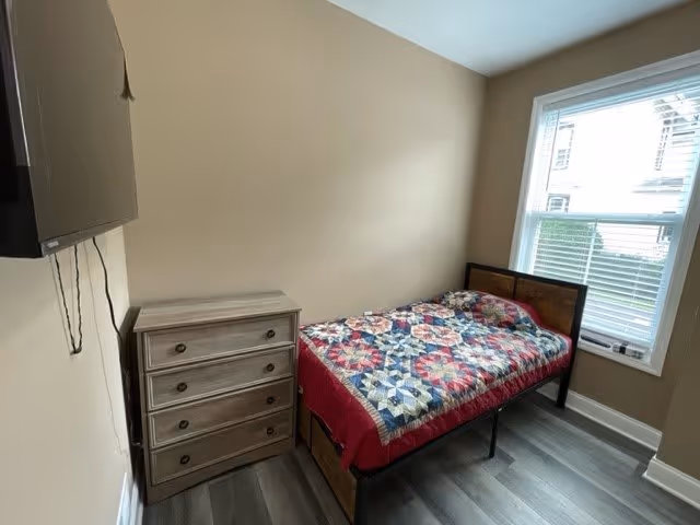 A small bedroom with a single bed covered with a colorful quilt, a wooden chest of drawers next to the bed, a wall-mounted TV, and a window letting in natural light.