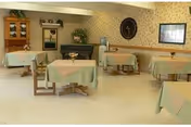 A dining room with several tables covered with light green tablecloths, each table having a small flower vase. The room has wallpaper with a floral pattern, a large wall clock, a framed picture, and a wooden cabinet with glass doors. The floor is light-colored and the room is well-lit.