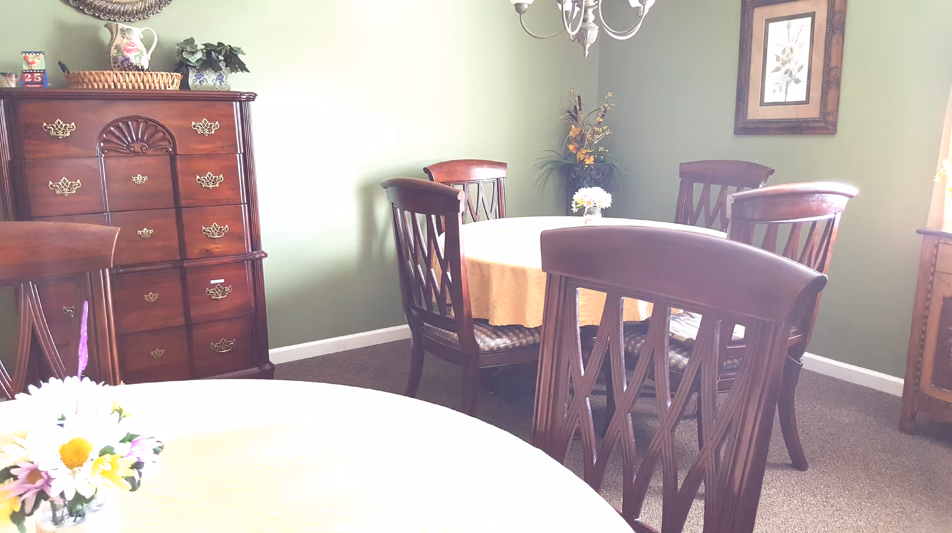A dining room with wooden chairs and tables covered with yellow tablecloths. There is a wooden chest of drawers against a green wall, decorated with a basket, a pitcher, and a small plant. A chandelier hangs from the ceiling, and a framed picture is on the wall. A small flower arrangement is on the table in the foreground.