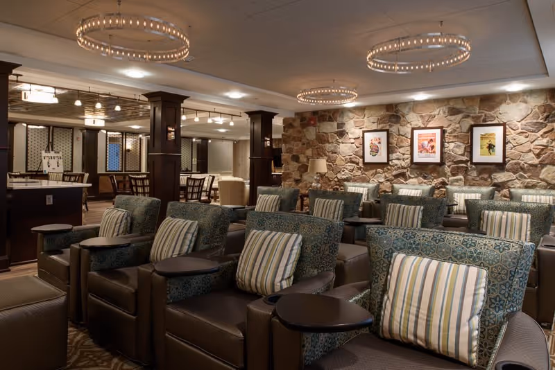 A cozy common area with multiple rows of comfortable armchairs featuring patterned upholstery and striped pillows, each equipped with small attached tables. The room has a warm ambiance with stone accent walls adorned with framed artwork, elegant circular chandeliers hanging from the ceiling, and dark wood columns and furniture. In the background, there are additional seating areas and tables.