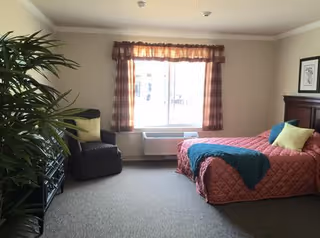 A cozy bedroom with a large window covered by plaid curtains. The room features a bed with a coral quilt, green and blue pillows, and a blue throw blanket. There is a dark armchair with a green pillow next to a tall leafy plant. A framed picture hangs on the wall above the bed, and an air conditioning unit is installed below the window.