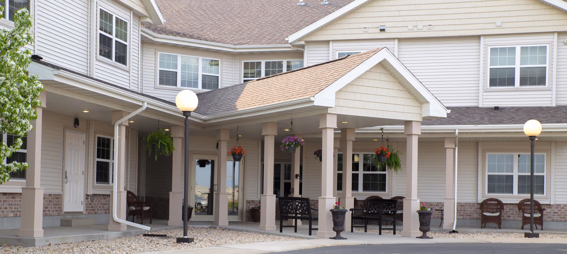 Exterior view of a senior living facility entrance with a covered porch supported by columns, outdoor seating, hanging flower baskets, and two lamp posts. The building has beige siding with brick accents and multiple windows.