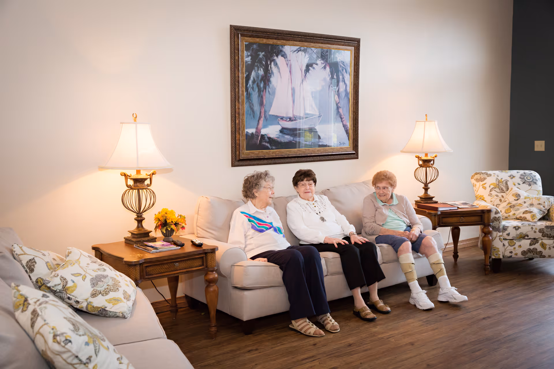 Three elderly women sitting on a beige sofa in a well-lit living room with wooden flooring. The room features two side tables with decorative lamps and a small flower arrangement, a framed painting of a sailboat on the wall, and a floral-patterned armchair.