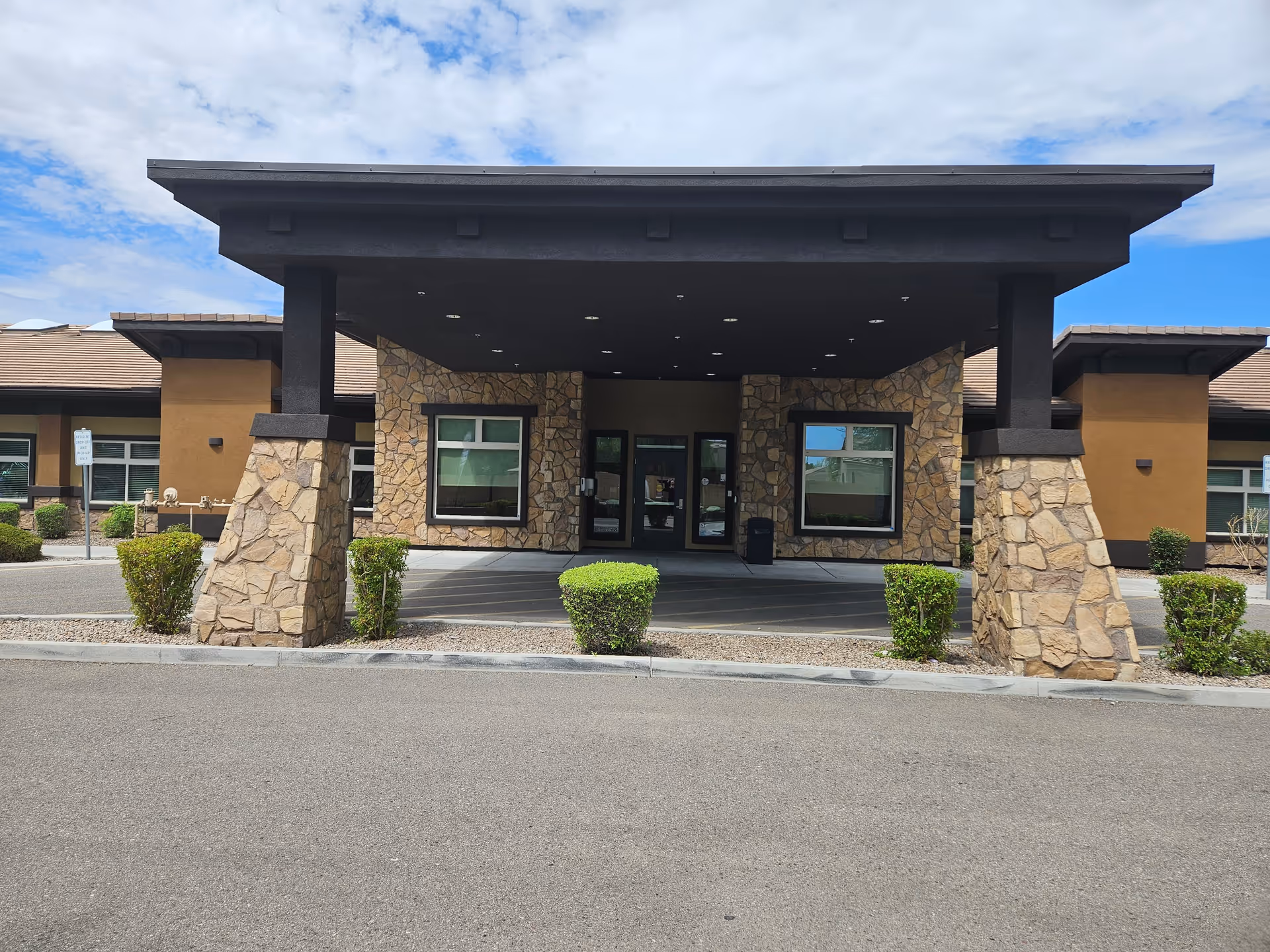 Front entrance of a building with stone pillars supporting a large overhang. The building has stone and tan colored walls with windows on either side of the glass double doors. Small green bushes line the front of the entrance area under a partly cloudy sky.