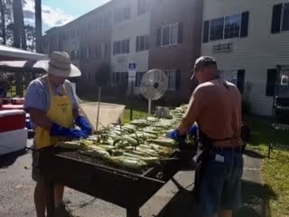 Two men wearing gloves and hats are grilling corn on an outdoor grill in front of a multi-story residential building on a sunny day.