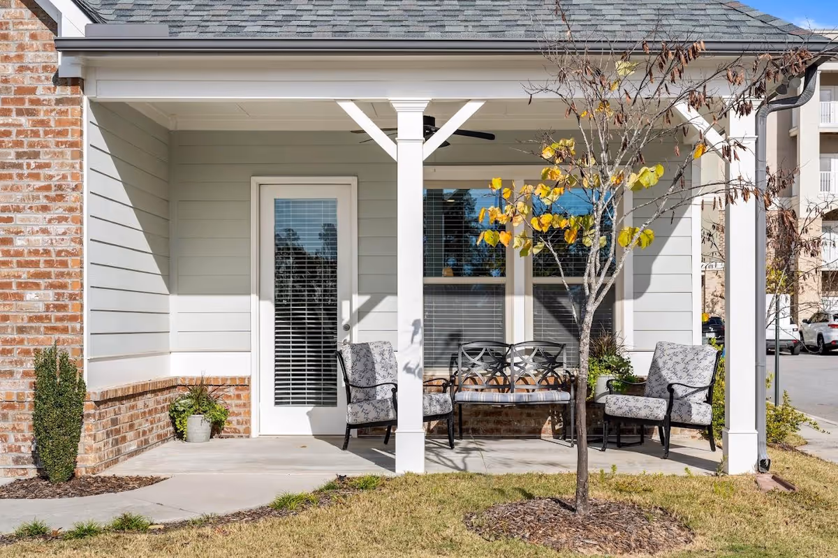 Outdoor covered patio area with two cushioned armchairs and a metal bench with cushions, a small tree with yellow and brown leaves in front, and a white door with window blinds leading inside the building.