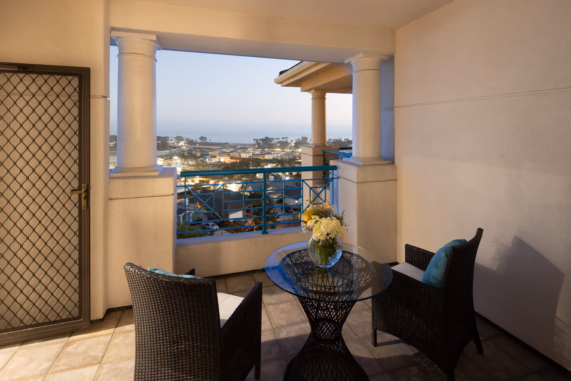 A covered balcony with two wicker chairs with cushions and a glass-top table holding a vase of flowers. The balcony overlooks a town with buildings and trees, and the ocean is visible in the distance under a clear sky at dusk.