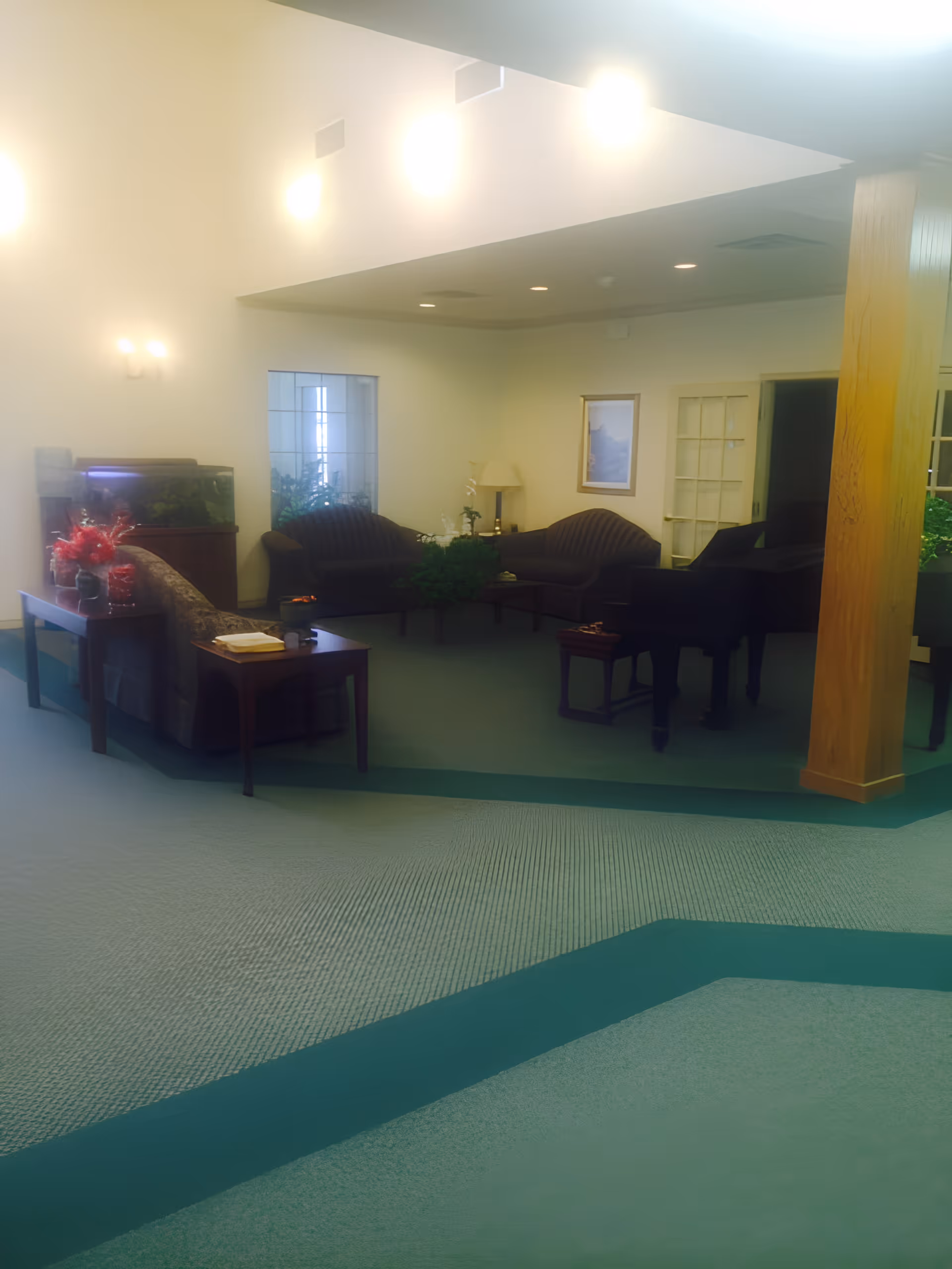 A spacious living room area in a senior living facility with green carpet and teal border. The room features several dark upholstered sofas arranged around a coffee table, a grand piano, a wooden pillar, a side table with a book and a cup, and an aquarium. The walls are light-colored with framed artwork and a lamp on a side table. Soft lighting fixtures illuminate the room.