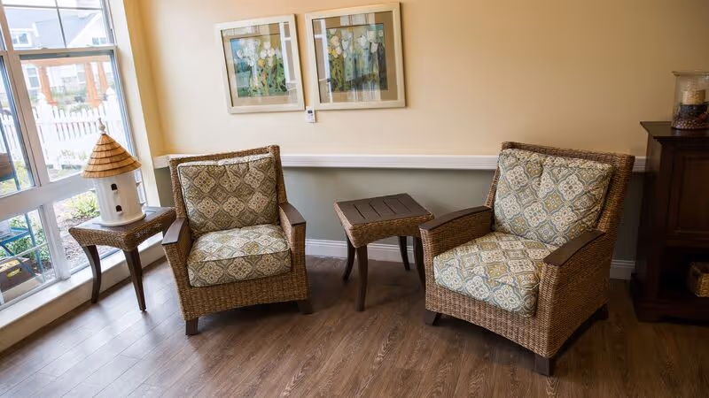 Two wicker armchairs with patterned cushions flank a small wooden table in a sunlit seating area by a large window with framed artwork on the wall.