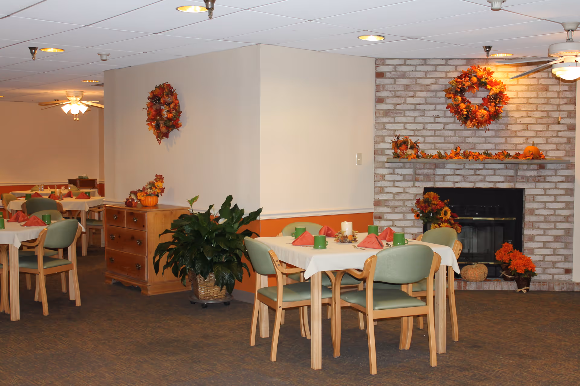 Dining room with tables set for a meal, green chairs, autumn decorations and a brick fireplace.
