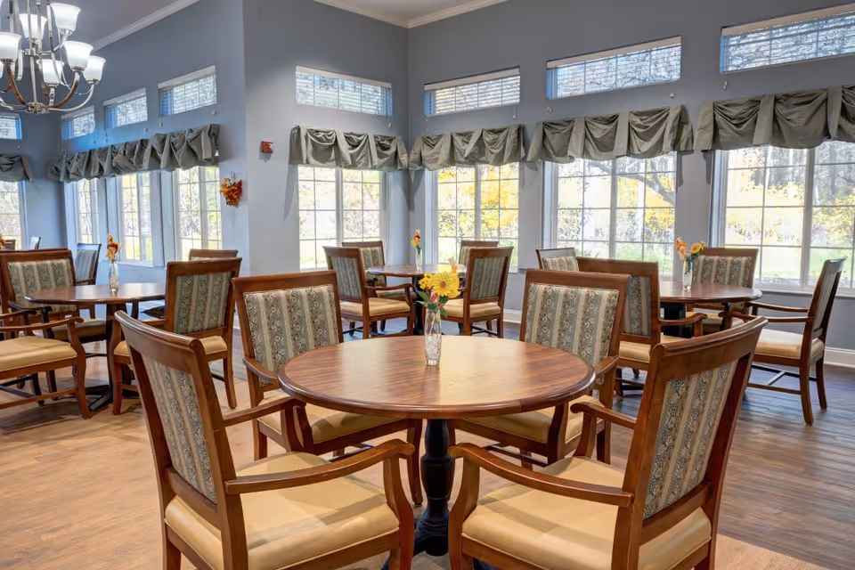 A bright dining room with multiple round wooden tables surrounded by cushioned wooden chairs. Each table has a small vase with flowers. Large windows with green valance curtains allow natural light to fill the room, and a chandelier hangs from the ceiling.