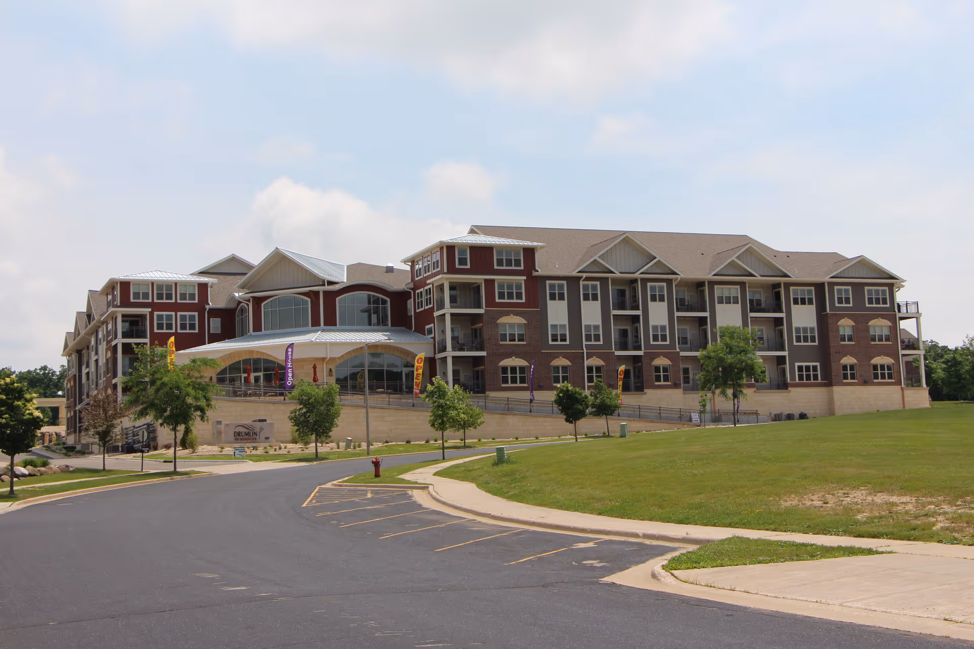 Exterior view of a large multi-story assisted living facility building with a mix of brick and siding, surrounded by a paved driveway, small trees, and grassy areas under a partly cloudy sky.