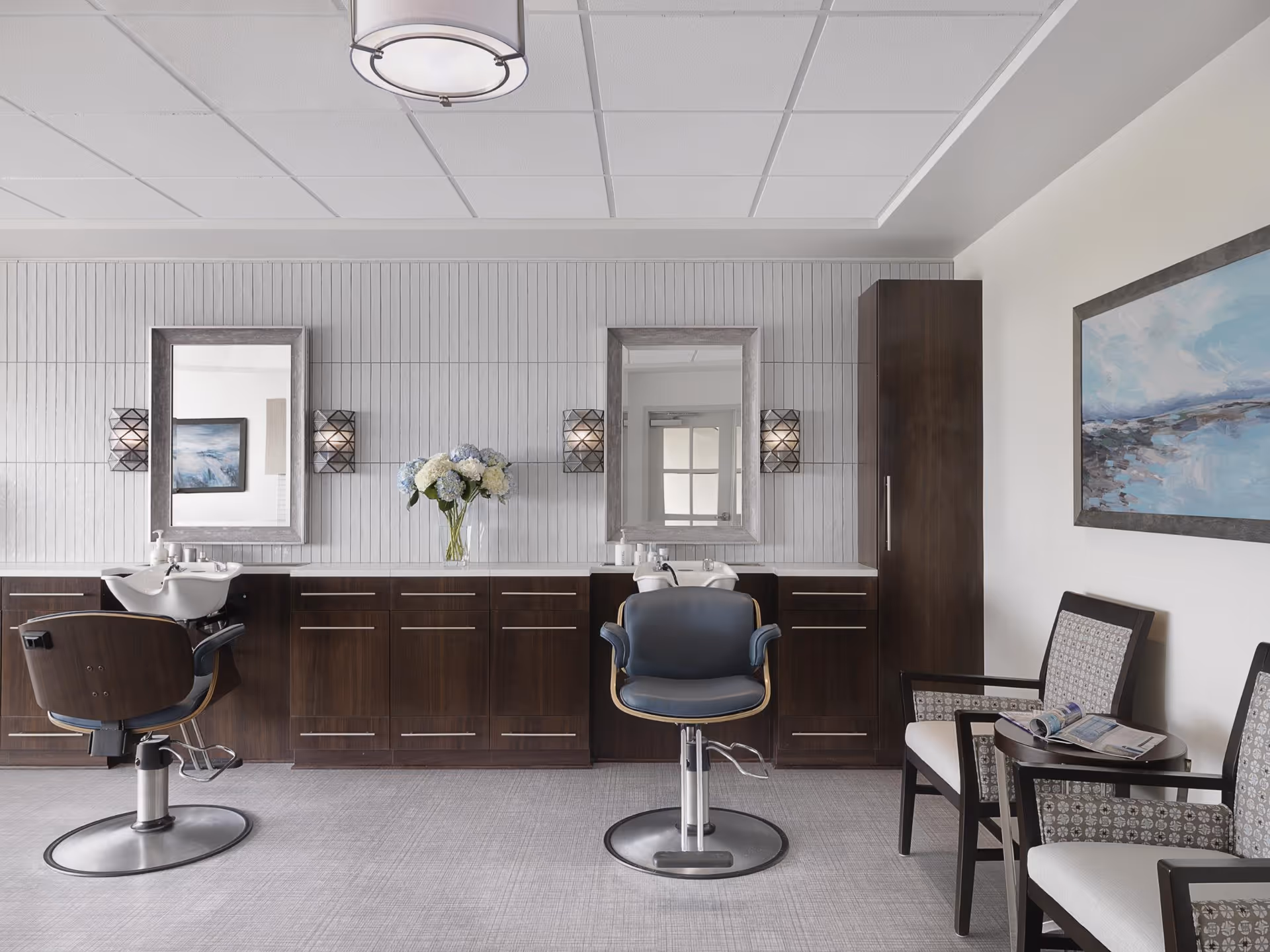 Interior view of a salon area in a senior living facility with two styling chairs in front of sinks and mirrors, dark wood cabinetry, a vase with flowers on the counter, and a seating area with two patterned chairs and a small table holding magazines. A framed painting hangs on the wall.