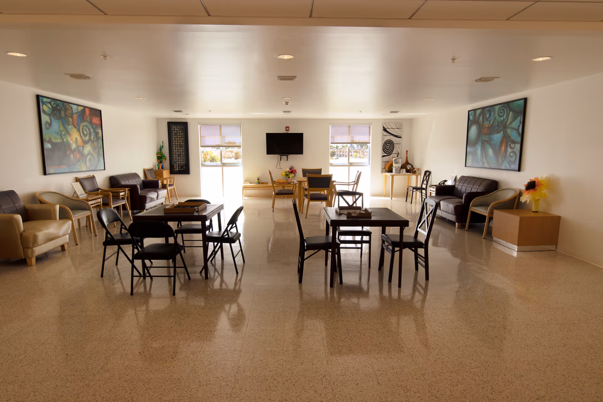 Spacious communal lounge with tables, chairs, sofas, wall art, and a TV at the far end.