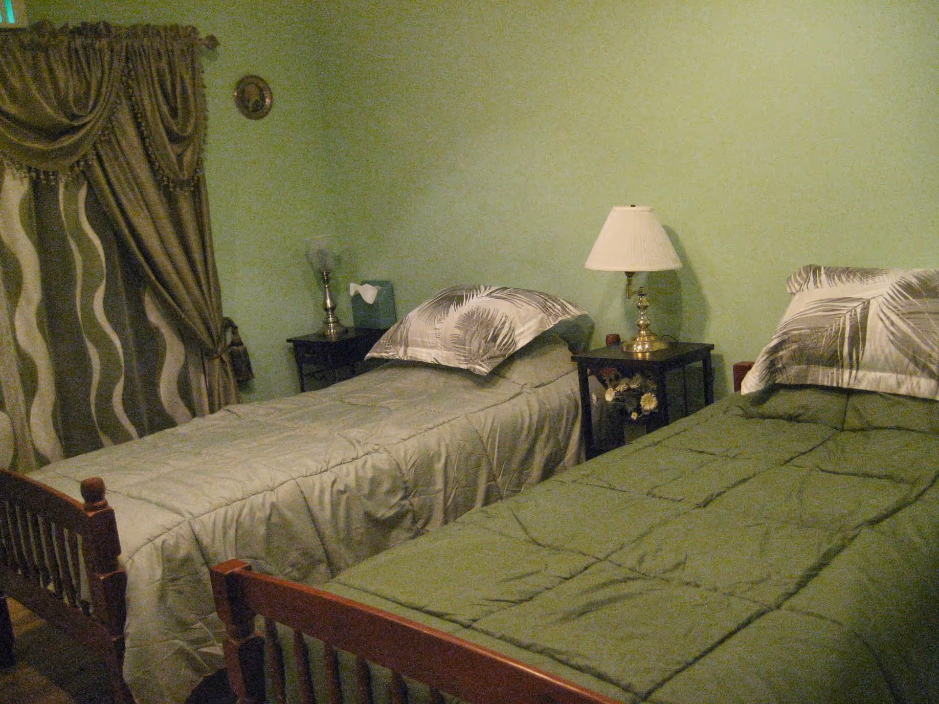 A bedroom with two single beds, each with green bedding and a pillow with a leaf pattern. Between the beds is a small dark wooden nightstand with a brass lamp and a small decorative item. The walls are painted light green, and there is a window with patterned curtains on the left side of the room.