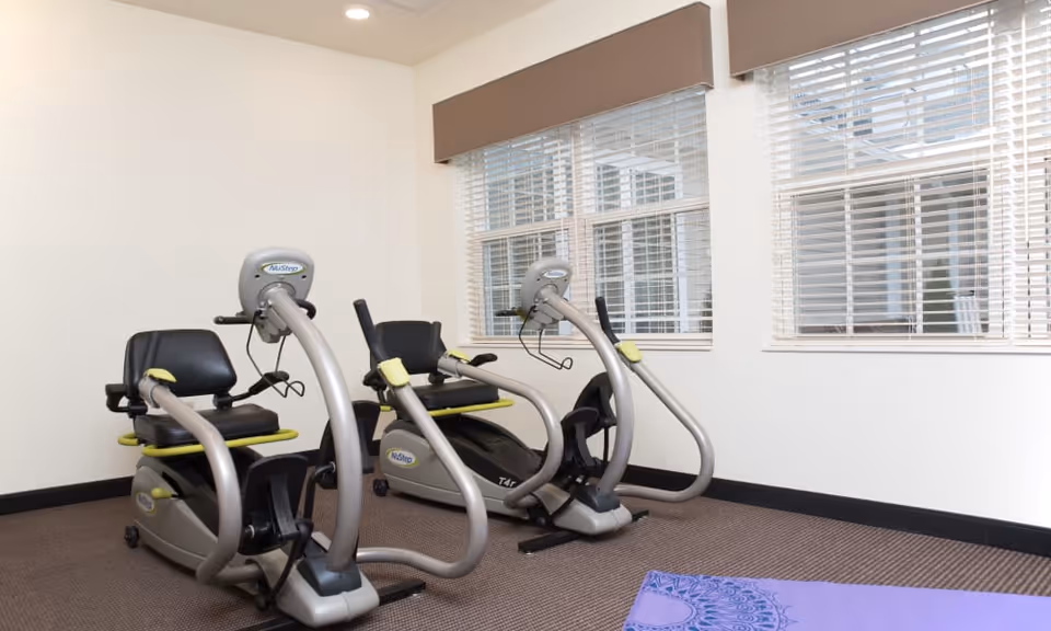 Two recumbent exercise bikes sit in a small fitness room with large windows and a purple yoga mat on the floor.