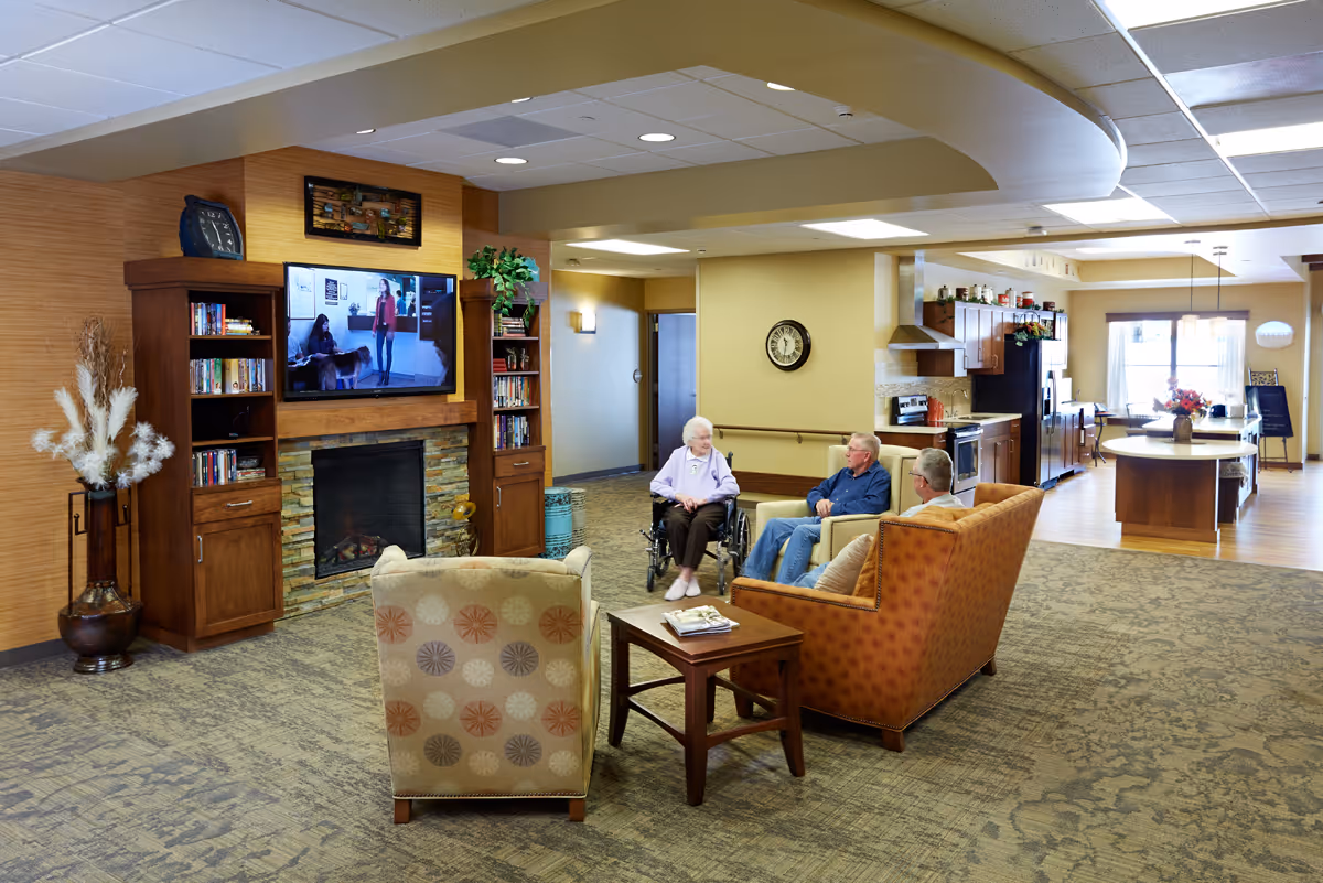 A cozy living room area in a senior living facility with three elderly people sitting and conversing. One person is in a wheelchair, and two others are seated on armchairs and a sofa. The room features a large flat-screen TV mounted above a stone fireplace, wooden bookshelves, and a decorative vase with dried plants. In the background, there is an open kitchen with wooden cabinets, a kitchen island, and a dining area with large windows letting in natural light.