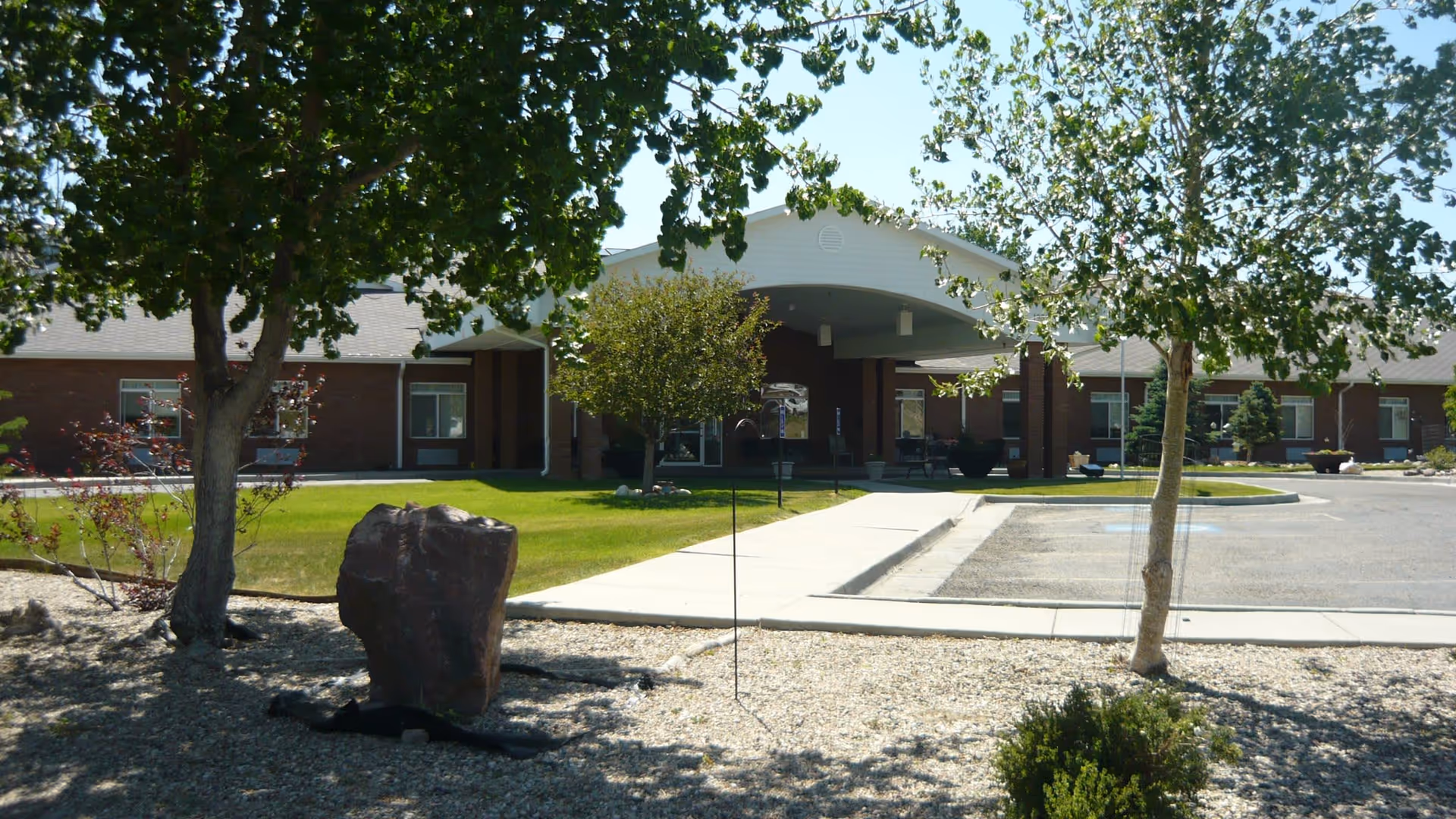 Exterior view of Garden Square Assisted Living of Casper showing a single-story brick building with a covered entrance, surrounded by green grass, trees, and a gravel area with a large rock.