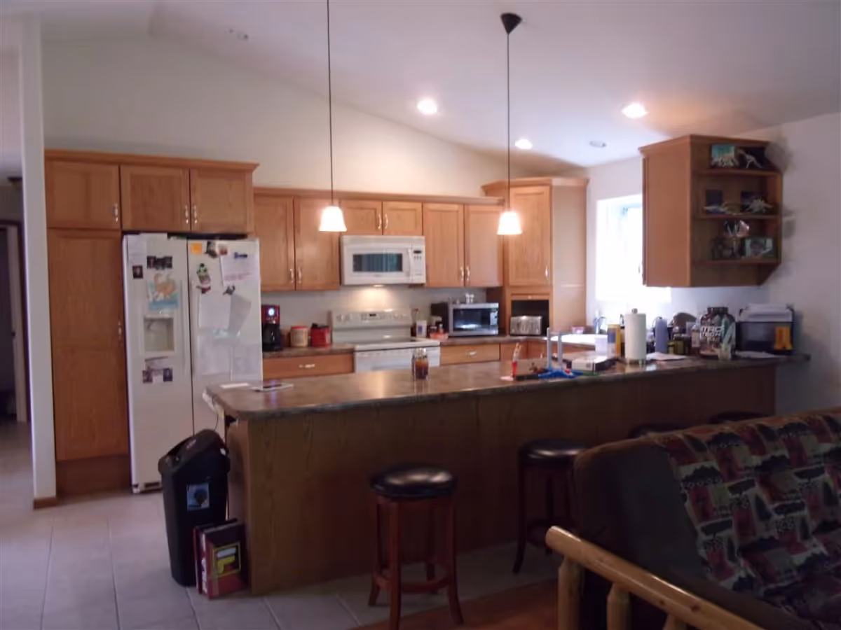 Open kitchen with wooden cabinets, a long island with stools, refrigerator and microwave, and a couch partially visible in the foreground.