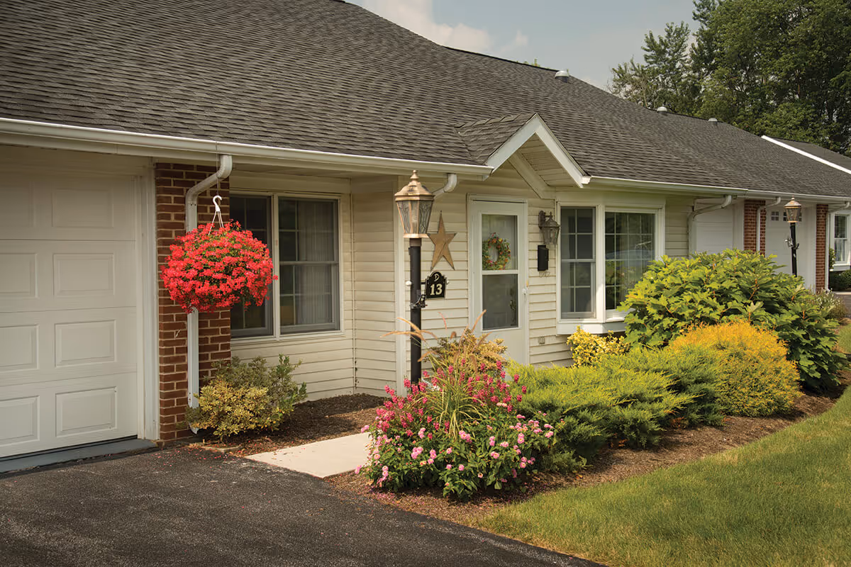 Exterior view of a single-story residential unit with beige siding and a dark shingled roof. The entrance features a white door with a wreath, a window to the right, and a hanging basket of red flowers to the left near a garage door. The front yard is landscaped with various green shrubs and flowering plants, and a black lamp post with the number 13 is visible near the walkway.