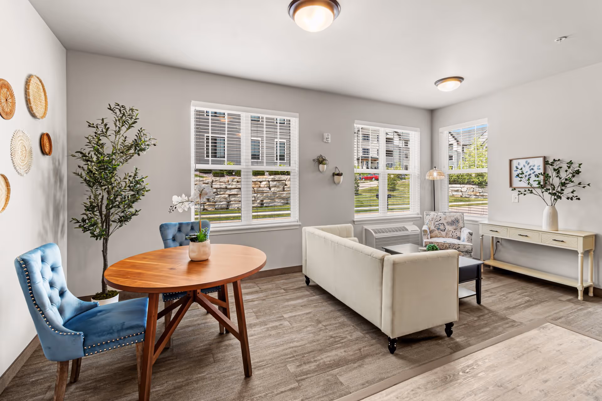 A bright and airy living room area with large windows letting in natural light. The room features a white sofa, a floral armchair, a wooden coffee table, and a long console table with a vase of greenery. There is also a small round wooden dining table with two blue upholstered chairs. Decorative woven wall hangings and a potted plant add warmth to the space.