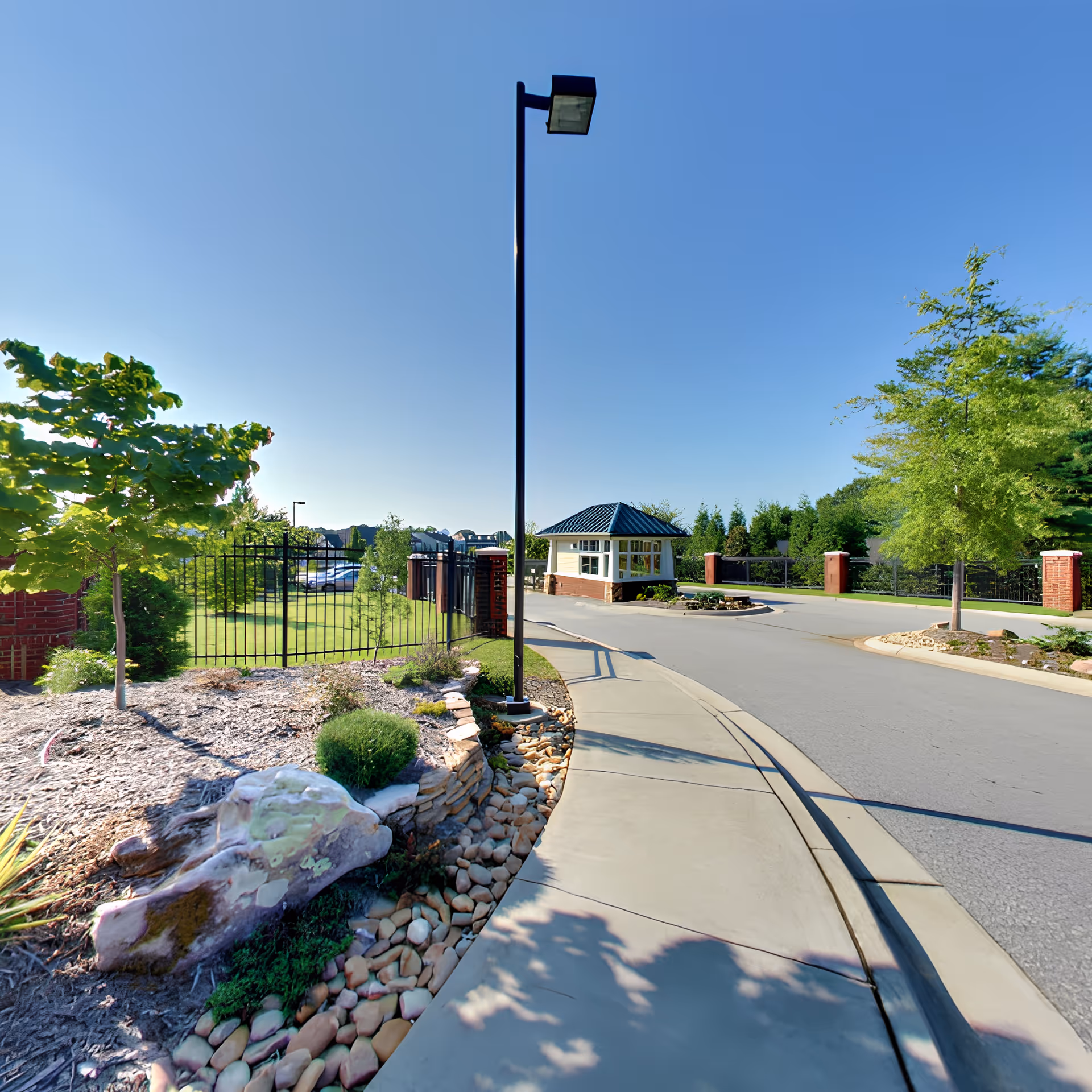 A paved road with a sidewalk curves past a small guardhouse with a blue roof at the entrance of a gated community. The area is landscaped with trees, bushes, rocks, and mulch under a clear blue sky.