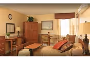 A cozy senior living bedroom with a bed covered in a beige quilt and decorative pillows, a wooden desk with a chair and lamp, a wooden armoire, a small round table with two chairs near a window with sheer curtains, and warm yellow walls.