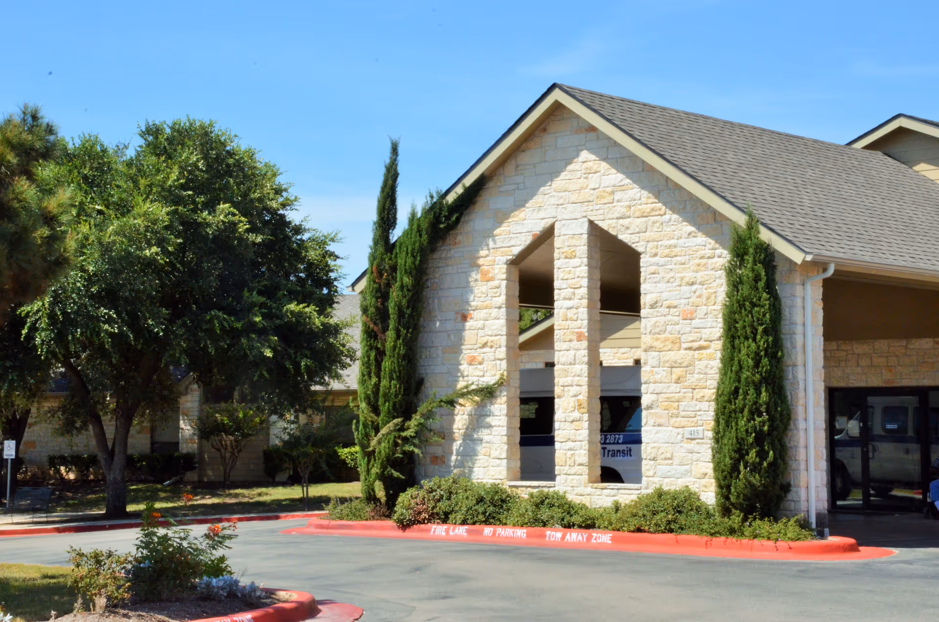 Exterior view of a stone building with a peaked roof, surrounded by trees and shrubs, with a driveway and red-painted curb marked as fire lane, no parking, tow away zone.