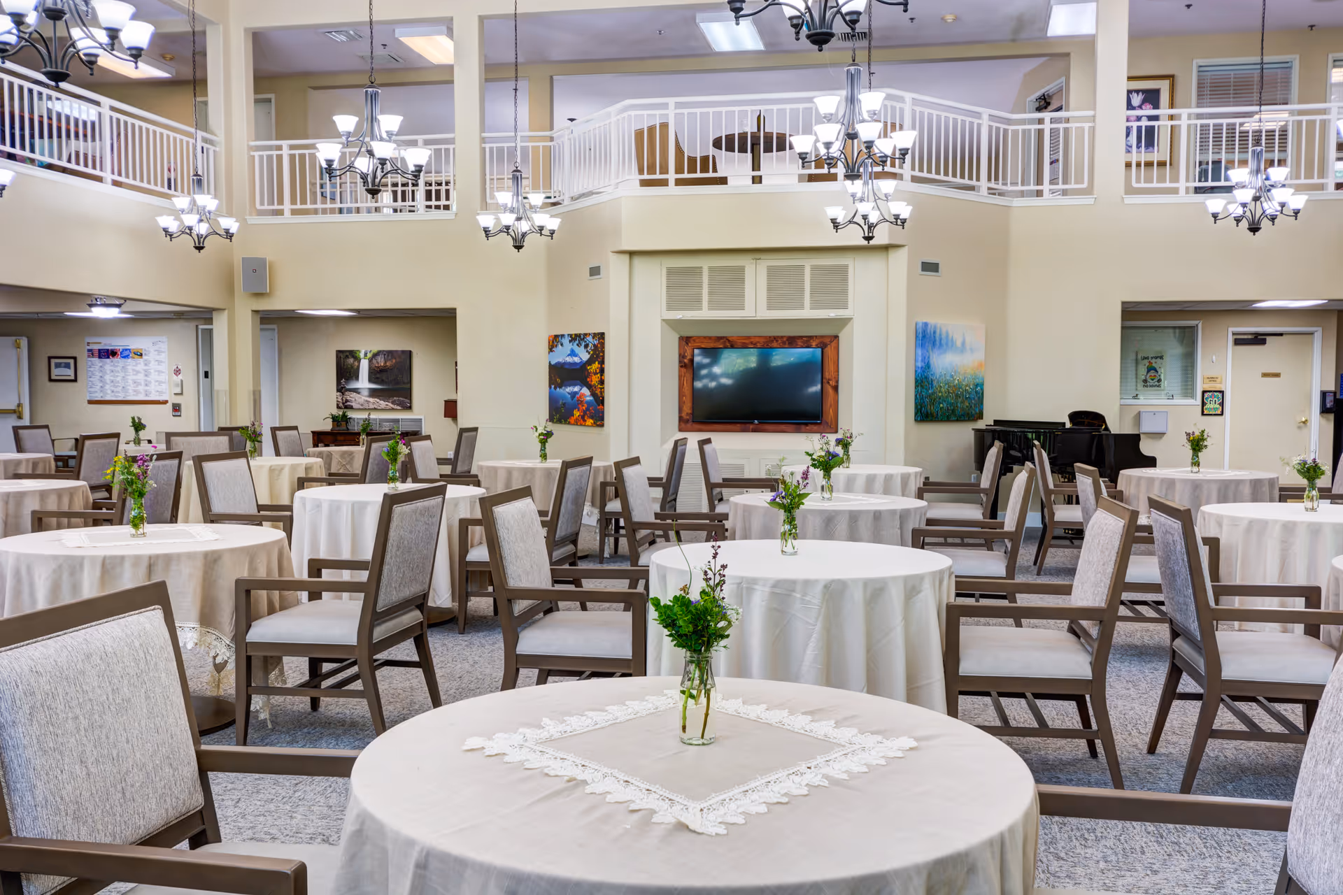 A spacious dining room with round tables covered in white tablecloths, each adorned with a small vase of flowers. The room features multiple chandeliers hanging from a high ceiling, a large flat-screen TV mounted on the far wall, and artwork displayed on the walls. There is a piano in the corner and a second-floor balcony overlooking the area.