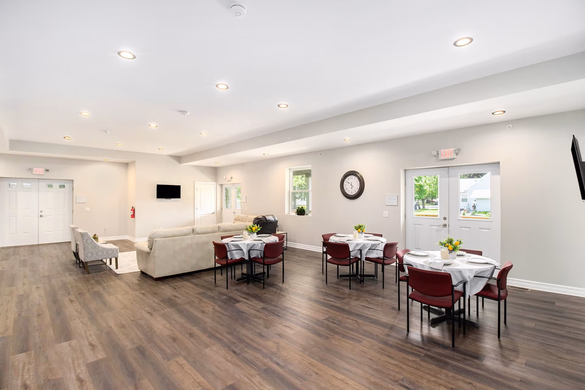 A spacious and bright common area in Evergreen Villas featuring several round dining tables with white tablecloths and red chairs, each table decorated with a small flower arrangement. There is a beige sectional sofa and two patterned armchairs arranged near a wall-mounted TV. The room has wood flooring, white walls, multiple windows, and recessed ceiling lights. Two exit doors are visible, along with a large wall clock.