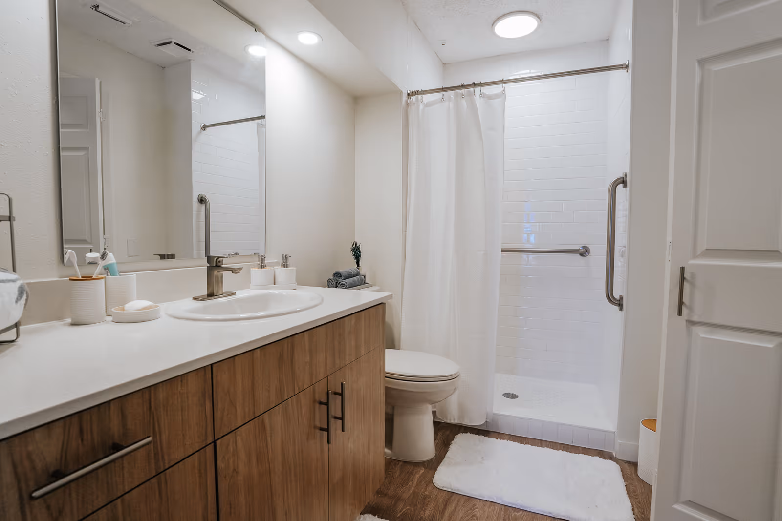 A clean and modern bathroom with a wooden vanity featuring a white countertop and sink. There is a large mirror above the sink, a toilet next to the vanity, and a walk-in shower with white subway tiles and a white shower curtain. The floor is wooden with white bath mats, and the bathroom has bright overhead lighting.