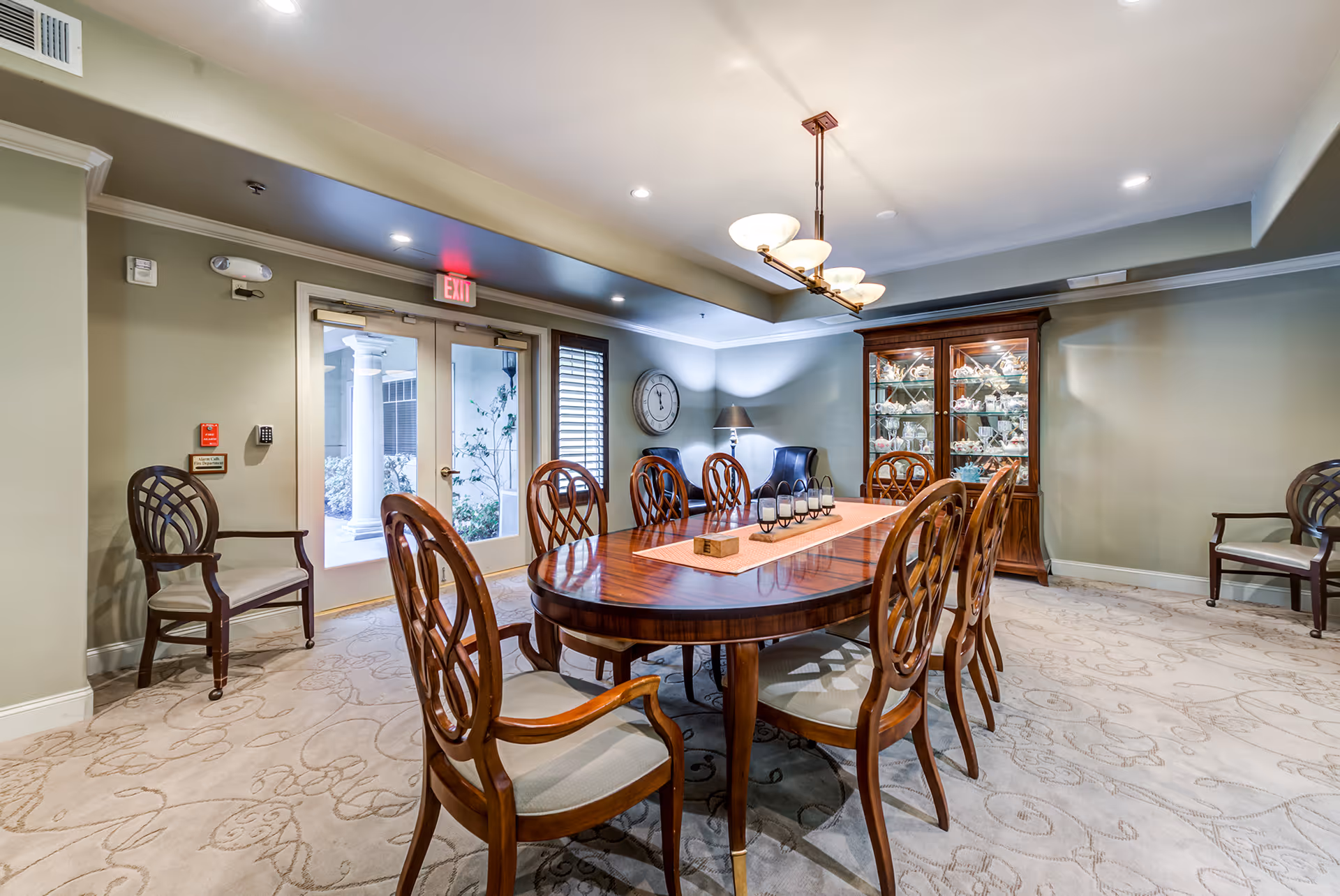 Well-lit dining room with a long wooden table surrounded by chairs, a china cabinet, and double glass doors.